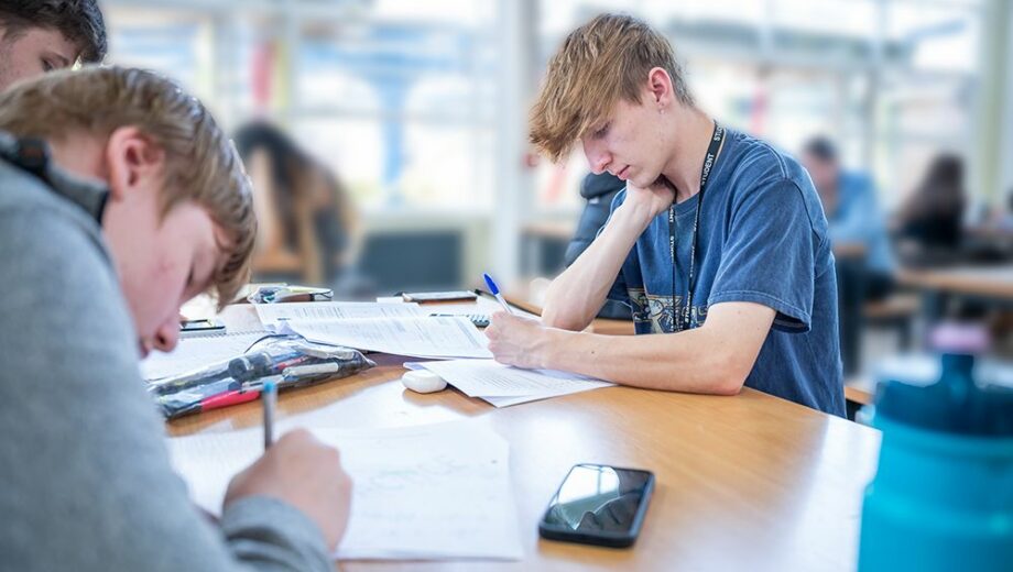 Male student sat at table, head down writing. a level courses