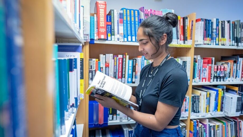 Female Student stood in library nook reading a book. a level courses
