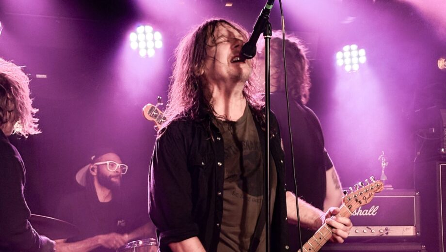 Photo of a male singing at a mic playing the guitar with lights around him