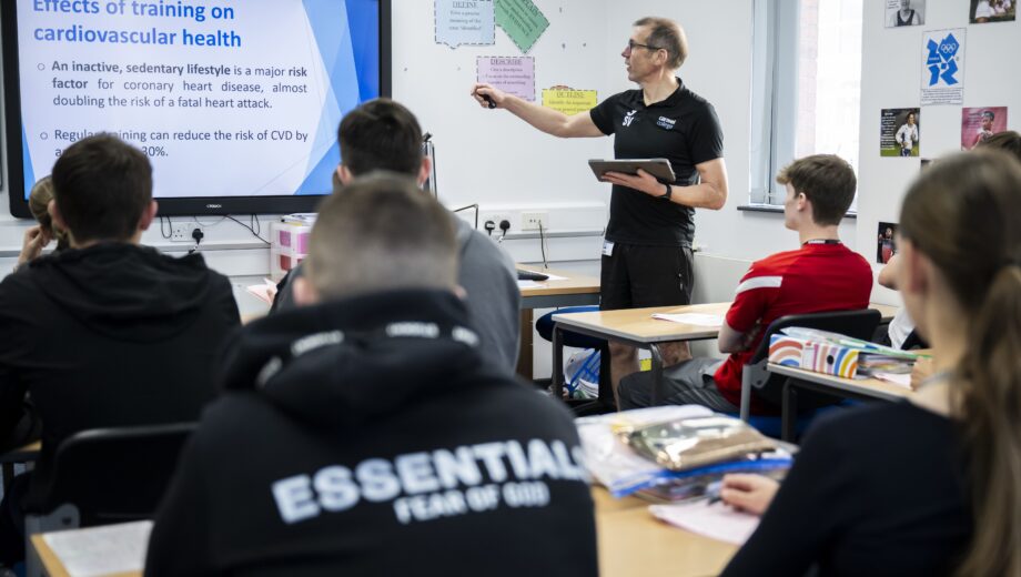 Male PE teacher stood at the white board in a PE classroom