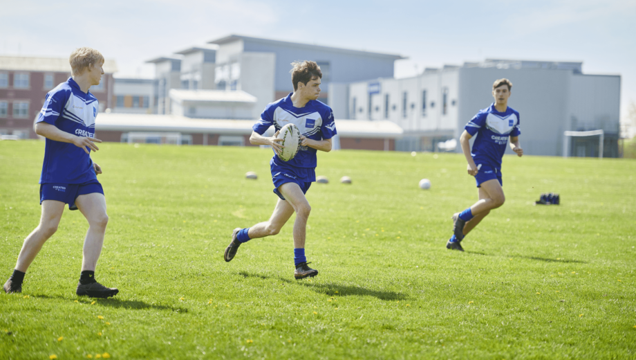 Three male students running on a field passing a rugby ball