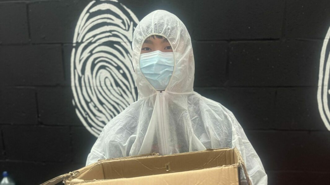 Chinese Student in a CSI bunny suit and blue paper mask holding a box against a black brick wall