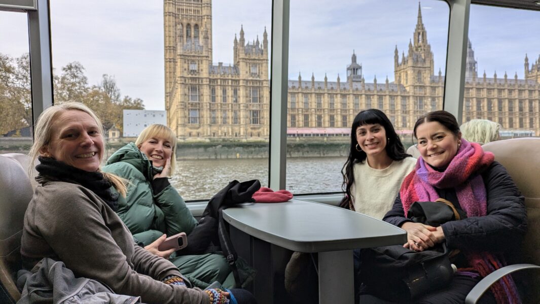 Carmel College Art and Design tutors sat around table on ferry smiling in front of the houses of parliament.