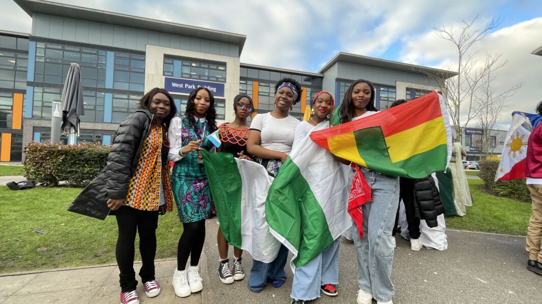 Group of female Carmel College African Caribbean society students dressed in cultural clothing hold Ghana flag.