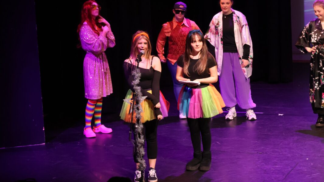 Christmas Panto fairies stand in front of microphone stand in rainbow tutus.