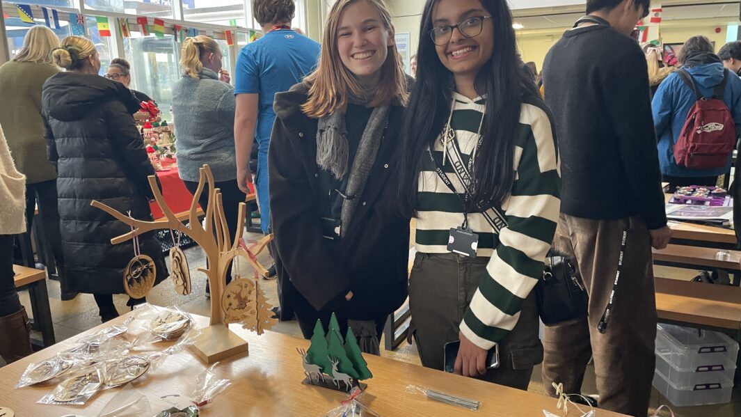 Two female students sand in front of hand made crafts stall at Carmel College's Christmas Market 2024.