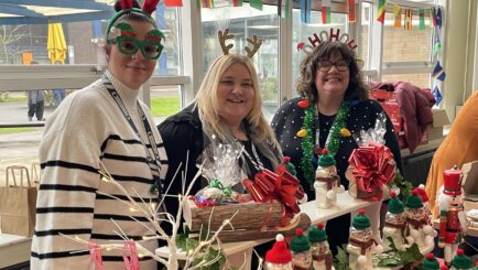 Three female Carmel College staff attend market stall wearing festive headbands.