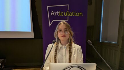 girl in white blazer, stood behind laptop giving a speech