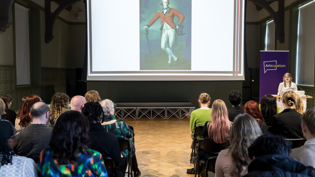 A room of people sat on chairs looking at a large screen with art work on.