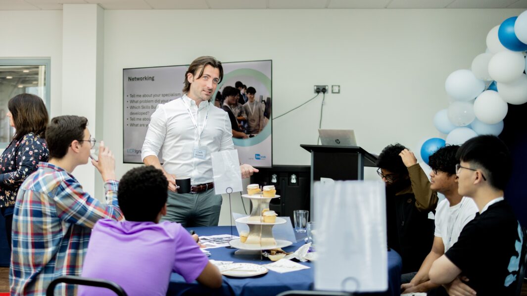 Male stood at the front of a round table talking to a group of male students