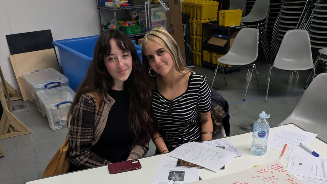 Two female students sat at a desk with artwork on a white table
