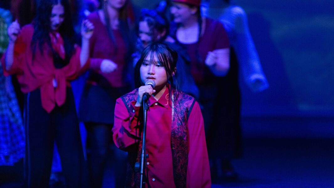 A large group of students dressed up on stage in the Wizard of Oz panto. A girl at the front wearing red is singing