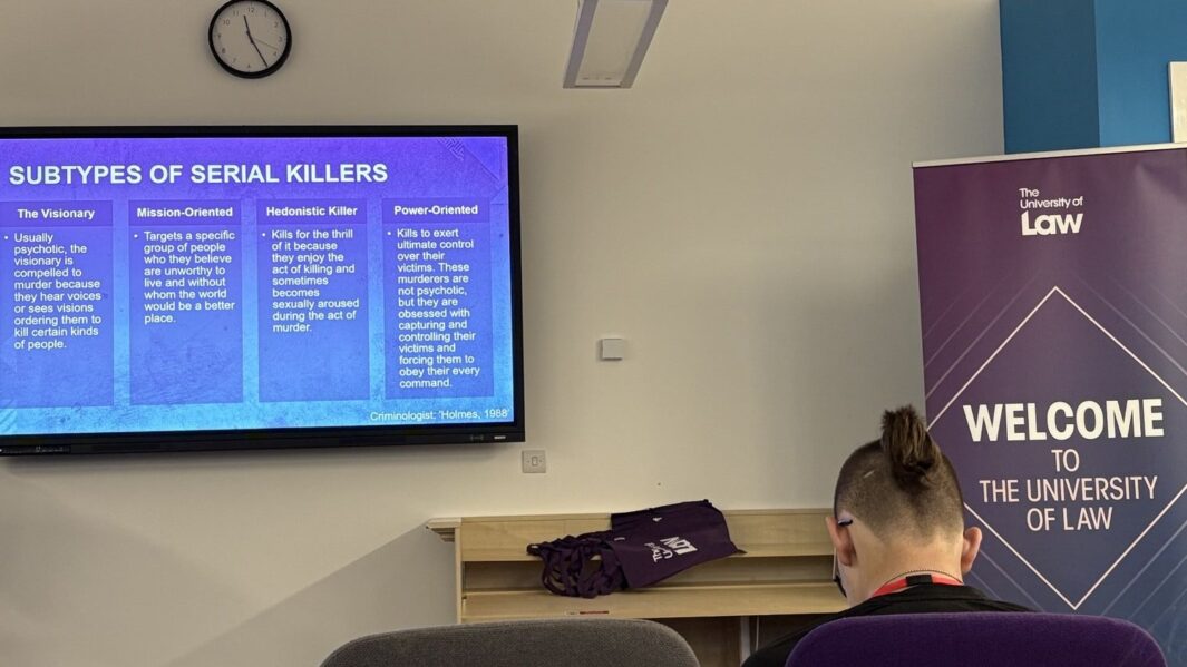 Image of the back of a man sat at a desk with a tv screen on the wall int he background