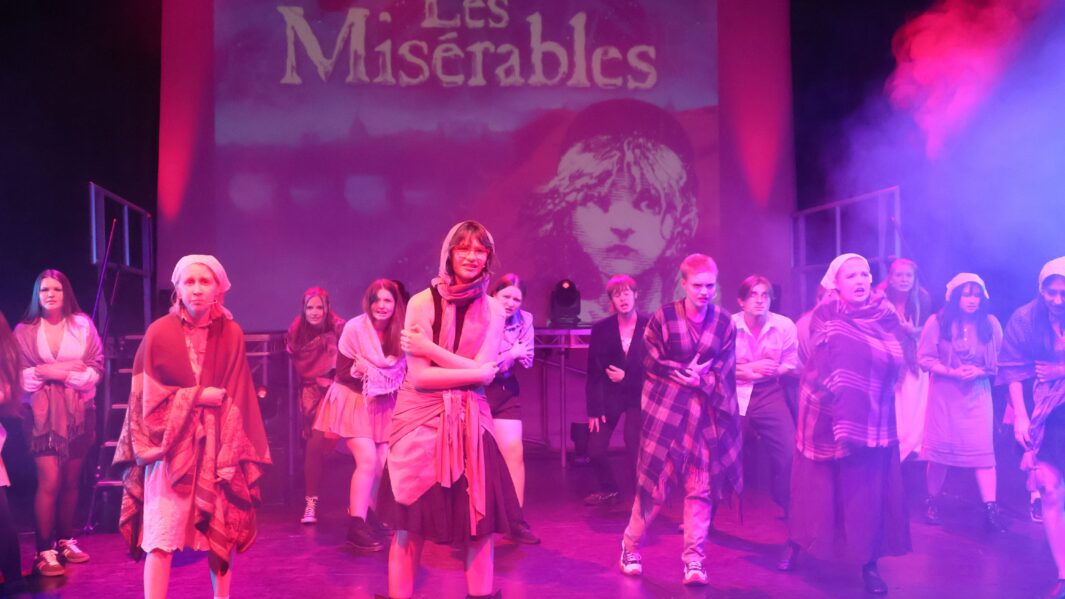 Large group of students (male and female) on stage in front of a les miserables backdrop