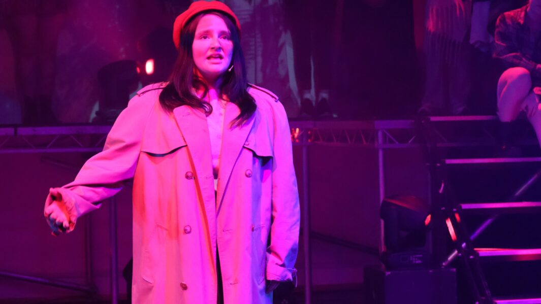 Girl on stage singing wearing a rain mac and hat with red lighting on her