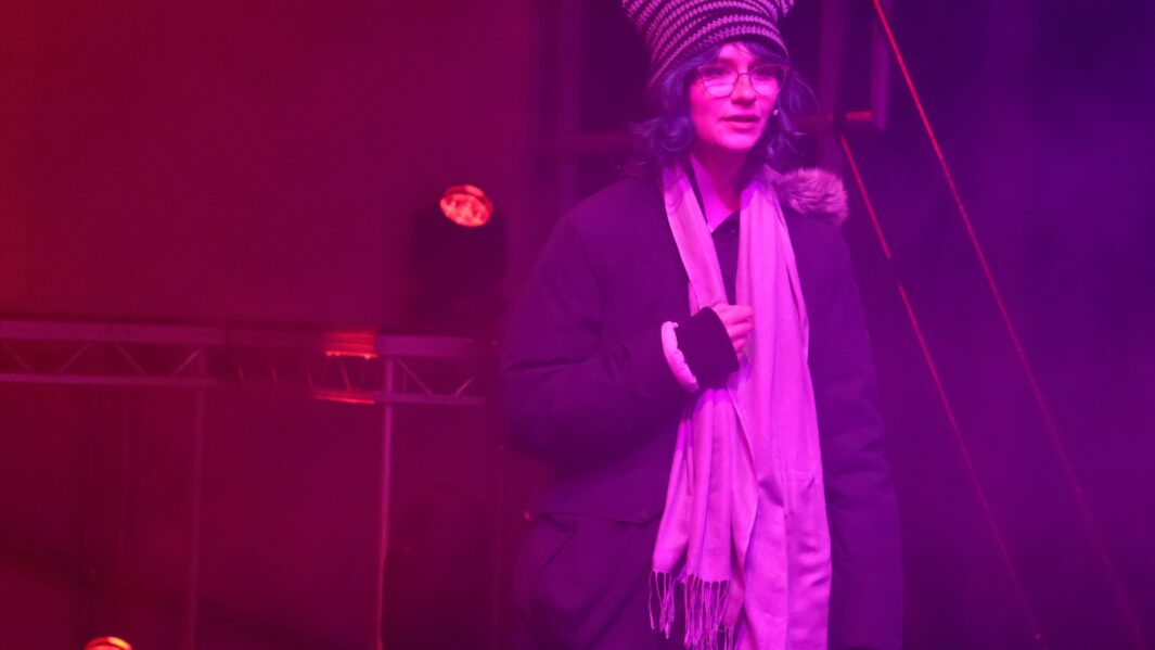 Girl with long brown hair singing on stage wearing a scarf and hat