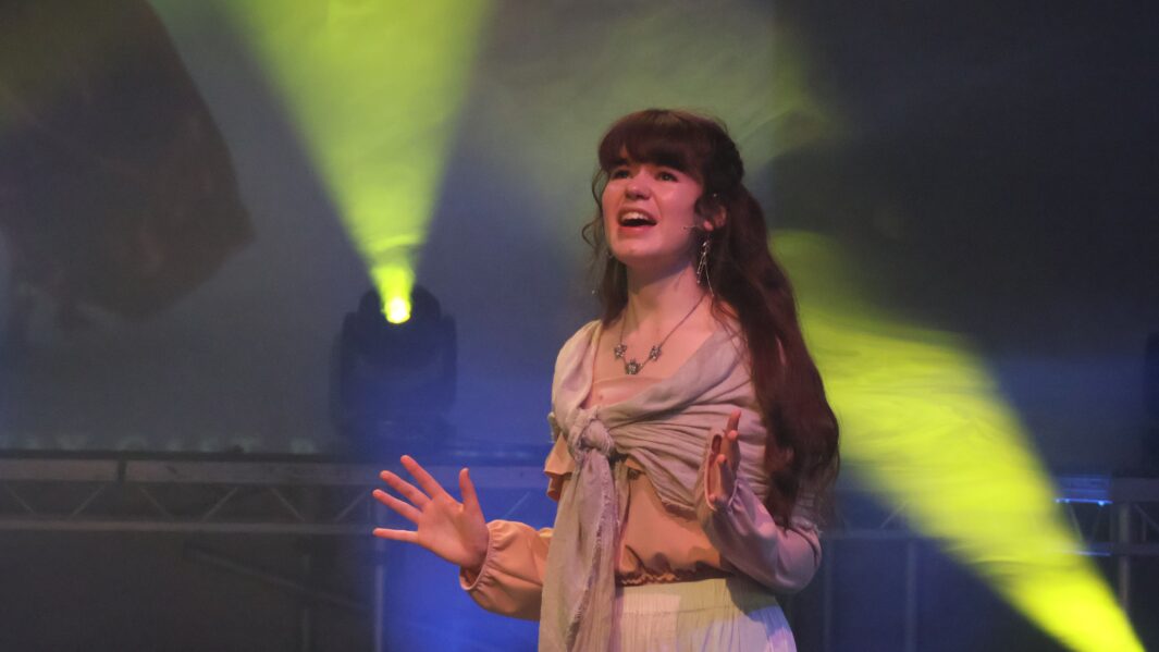Girl with long brown hair singing on stage wearing a shawl